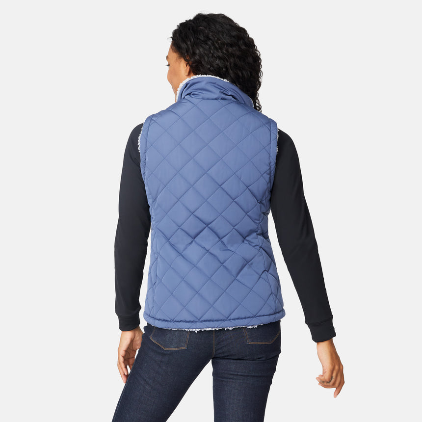 [Angelique is 5'10" wearing size S.] Women’s Free Country blue diamond-quilted vest with zip-front closure—reverses to cozy sherpa for added warmth and texture. Styled over a black long-sleeve shirt with one hand in pocket, set against a clean white background.