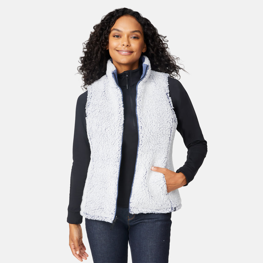 [Angelique is 5'10" wearing size S.] Women’s Free Country blue diamond-quilted vest with zip-front closure—reverses to cozy sherpa for added warmth and texture. Styled over a black long-sleeve shirt with one hand in pocket, set against a clean white background.
