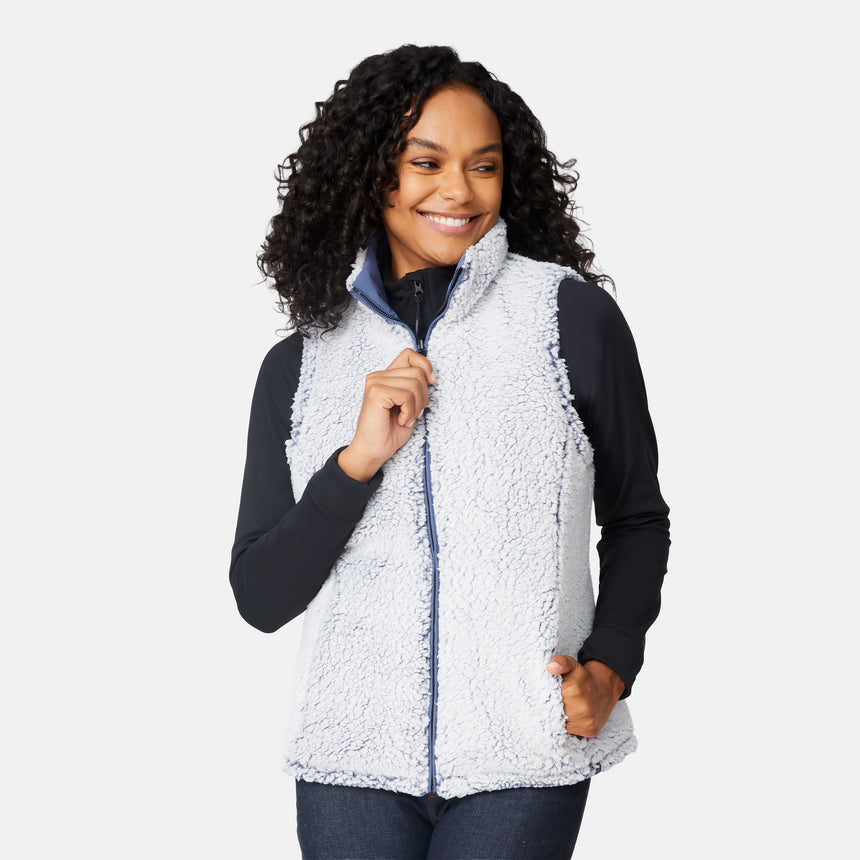 [Angelique is 5'10" wearing size S.] Women’s Free Country blue diamond-quilted vest with zip-front closure—reverses to cozy sherpa for added warmth and texture. Styled over a black long-sleeve shirt with one hand in pocket, set against a clean white background.