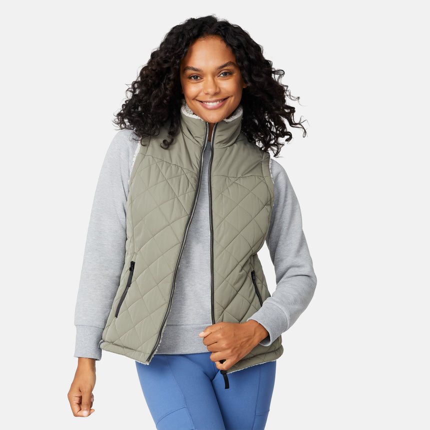 [Angelique is 5'10" wearing size S.] Women’s Free Country light green quilted vest with diamond pattern and black zippered pockets—reverses to cozy sherpa for added warmth and texture. Styled over a gray long-sleeve shirt and blue pants for a relaxed, transitional-weather look.