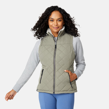 [Angelique is 5'10" wearing size S.] Women’s Free Country light green quilted vest with diamond pattern and black zippered pockets—reverses to cozy sherpa for added warmth and texture. Styled over a gray long-sleeve shirt and blue pants for a relaxed, transitional-weather look.