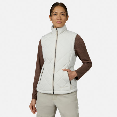A woman stands with one hand in her pocket against a plain white background, showcasing lightweight warmth and effortless style in the Free Country Women’s Stratus Lite II Reversible Vest in Bisque, crafted from recycled polyester and worn over a brown long-sleeve shirt and light pants.