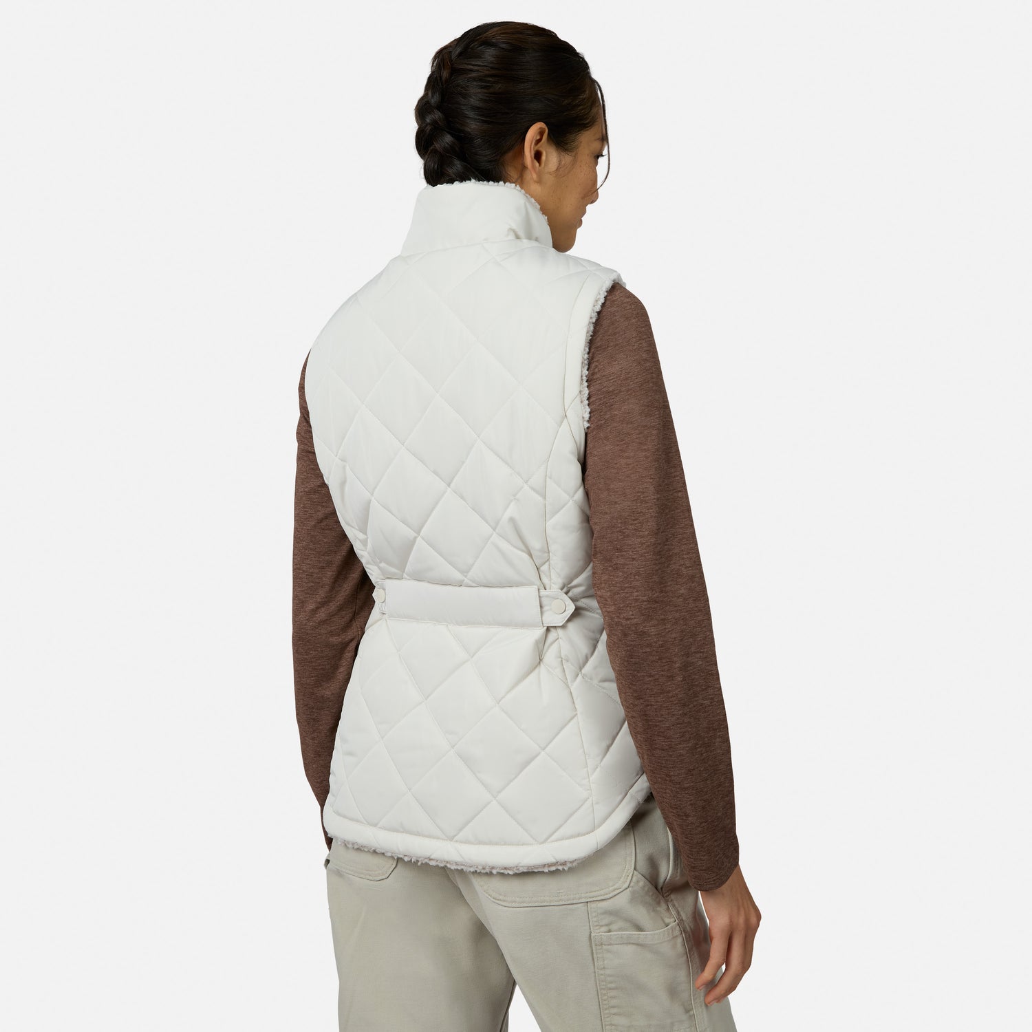 A person with braided hair stands with their back to the camera against a plain light background, wearing the Free Country Women’s Stratus Lite II Reversible Vest in white—crafted from recycled polyester—layered over a long-sleeve brown shirt and beige pants. This ensemble delivers lightweight warmth while maintaining an effortlessly stylish appearance.