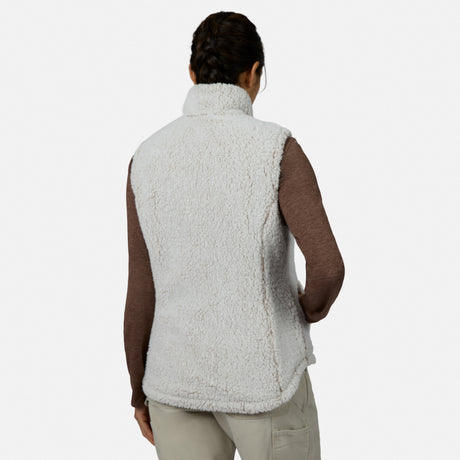 A person with braided hair stands with their back to the camera against a plain background, wearing the Free Country Women’s Stratus Lite II Reversible Vest—an eco-friendly, white, fuzzy sherpa outer layer made from recycled polyester—over a long-sleeve brown shirt and light pants. This stylish vest provides lightweight warmth for any occasion.