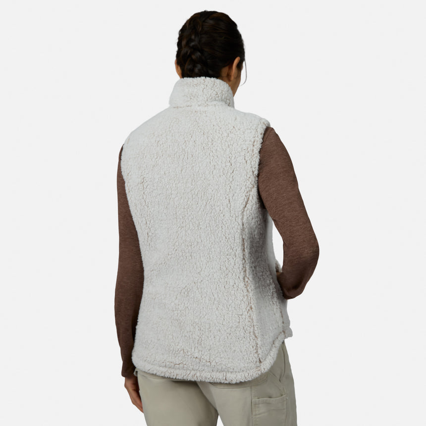 A person with braided hair stands with their back to the camera against a plain background, wearing the Free Country Women’s Stratus Lite II Reversible Vest—an eco-friendly, white, fuzzy sherpa outer layer made from recycled polyester—over a long-sleeve brown shirt and light pants. This stylish vest provides lightweight warmth for any occasion.