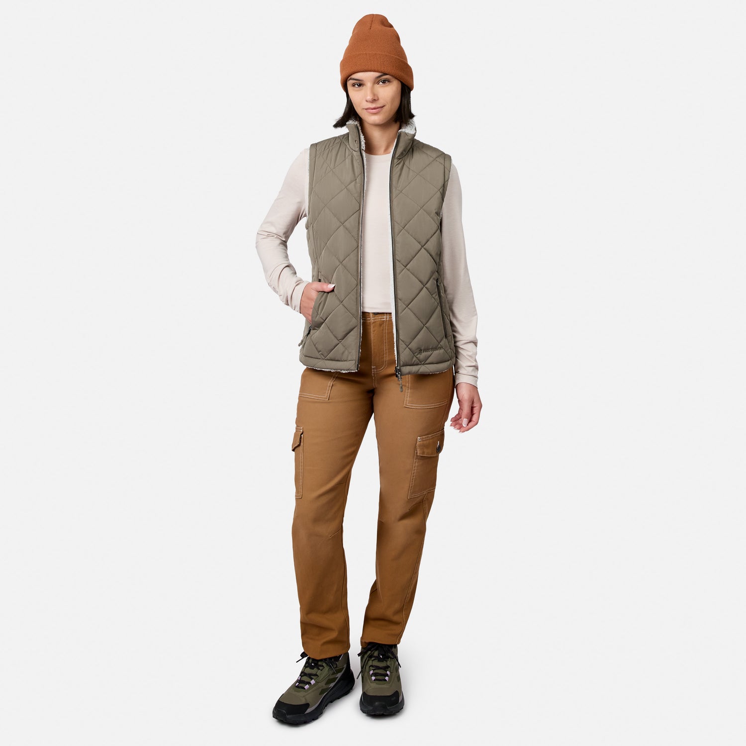 A person stands facing forward against a plain white background, wearing a brown beanie, the Free Country Women’s Stratus Lite II Reversible Vest—a quilted olive vest made from recycled polyester for lightweight warmth—a light long-sleeve shirt, brown cargo pants, and hiking boots.