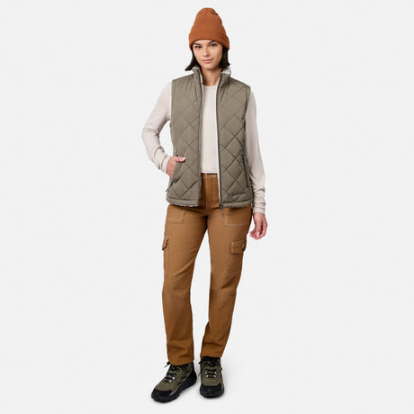 A person stands facing forward against a plain white background, wearing a brown beanie, the Free Country Women’s Stratus Lite II Reversible Vest—a quilted olive vest made from recycled polyester for lightweight warmth—a light long-sleeve shirt, brown cargo pants, and hiking boots.