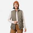 A smiling person stands against a plain white background, effortlessly stylish in a brown beanie, light long-sleeve shirt, brown pants, and the Women’s Stratus Lite II Reversible Vest by Free Country in Olivine—showcasing lightweight warmth from recycled polyester.