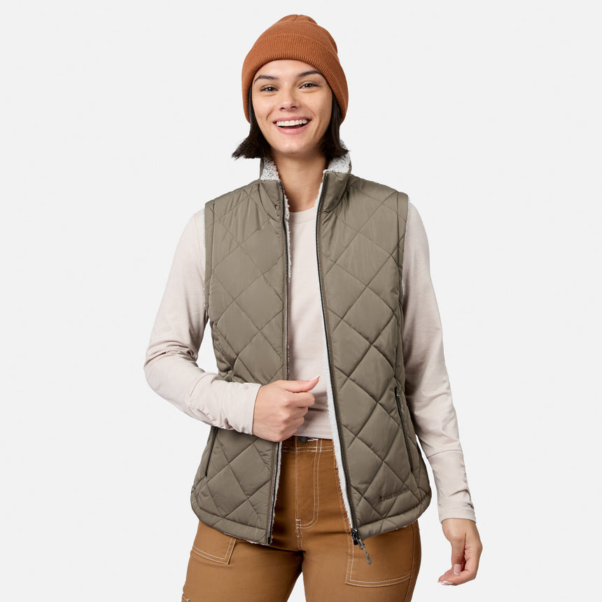 A smiling person stands against a plain white background, effortlessly stylish in a brown beanie, light long-sleeve shirt, brown pants, and the Women’s Stratus Lite II Reversible Vest by Free Country in Olivine—showcasing lightweight warmth from recycled polyester.