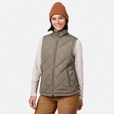 A woman smiles at the camera against a plain white background, wearing the Free Country Women’s Stratus Lite II Reversible Vest in tan, layered over a long-sleeve shirt, with brown pants and an orange knit beanie.
