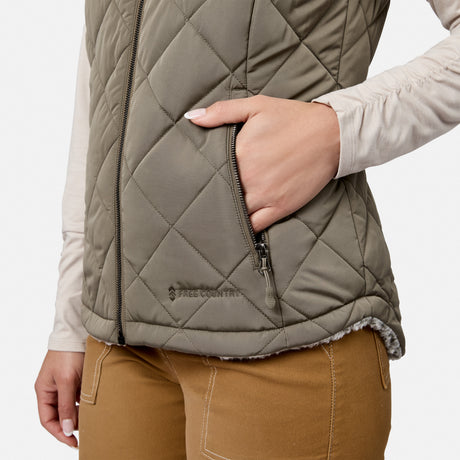 A woman wears the Free Country Women’s Stratus Lite II Reversible Vest in quilted olive green, featuring a visible zipper pocket, paired with tan pants. Designed for lightweight warmth, she rests one hand inside the pocket.