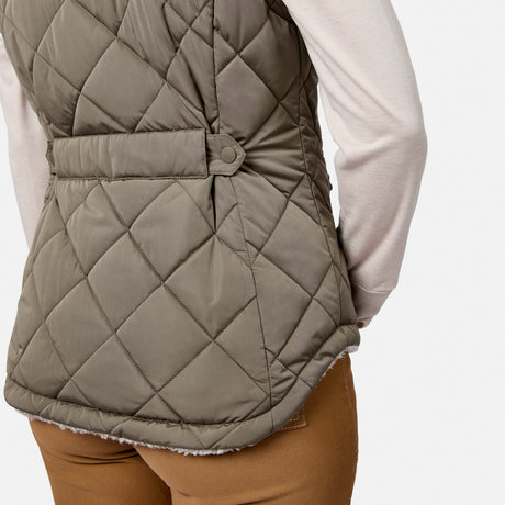 A person is seen from the back and partial left side, wearing the Free Country Women’s Stratus Lite II Reversible Vest in quilted olive green, crafted from recycled polyester with a belted waist. Layered over a light long-sleeve shirt and brown pants, this versatile piece highlights its lightweight warmth and eco-friendly design.