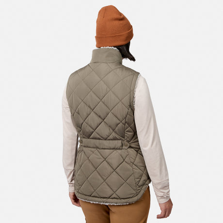 A person is standing with their back to the camera against a plain white background, wearing a brown beanie, brown pants, a light long-sleeve shirt, and the Free Country Women’s Stratus Lite II Reversible Vest—a beige vest made from recycled polyester for lightweight warmth.