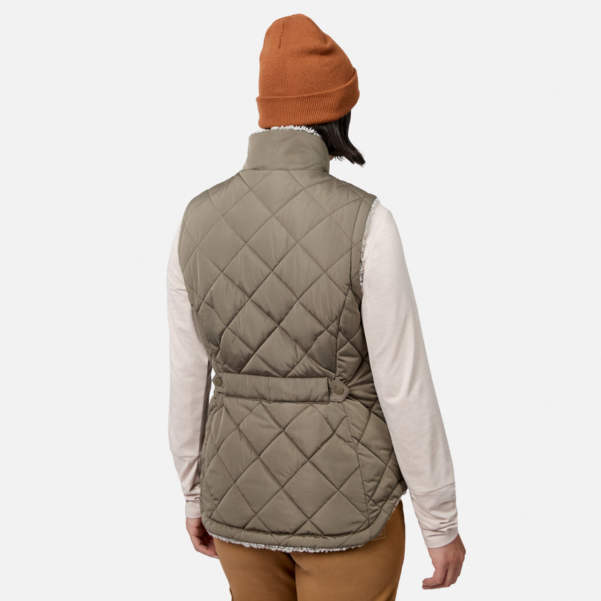 A person is standing with their back to the camera against a plain white background, wearing a brown beanie, brown pants, a light long-sleeve shirt, and the Free Country Women’s Stratus Lite II Reversible Vest—a beige vest made from recycled polyester for lightweight warmth.