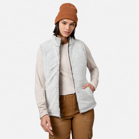 A woman stands against a plain white background with one hand in her pocket, showcasing effortless style and lightweight warmth in the Free Country Women’s Stratus Lite II Reversible Vest, a light gray fleece piece made from recycled polyester. She pairs it with a beige long-sleeve shirt, brown beanie, and brown pants.
