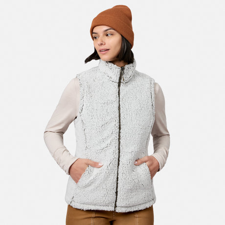 A woman stands against a plain background with her hands in the pockets of her Free Country Women’s Stratus Lite II Reversible Vest. She pairs the light gray vest—crafted from recycled polyester for lightweight warmth and effortless style—with a brown beanie, a beige long-sleeve shirt, and brown pants, looking off to the side.