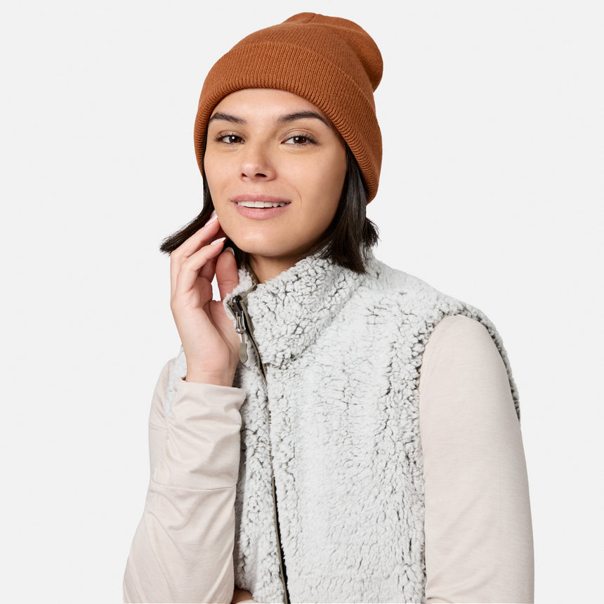 Wearing the Free Country Women’s Stratus Lite II Reversible Vest—crafted from recycled polyester for lightweight warmth—along with a brown knit beanie and a long-sleeve beige shirt, a woman smiles gently and touches her face with one hand against a plain, light background.