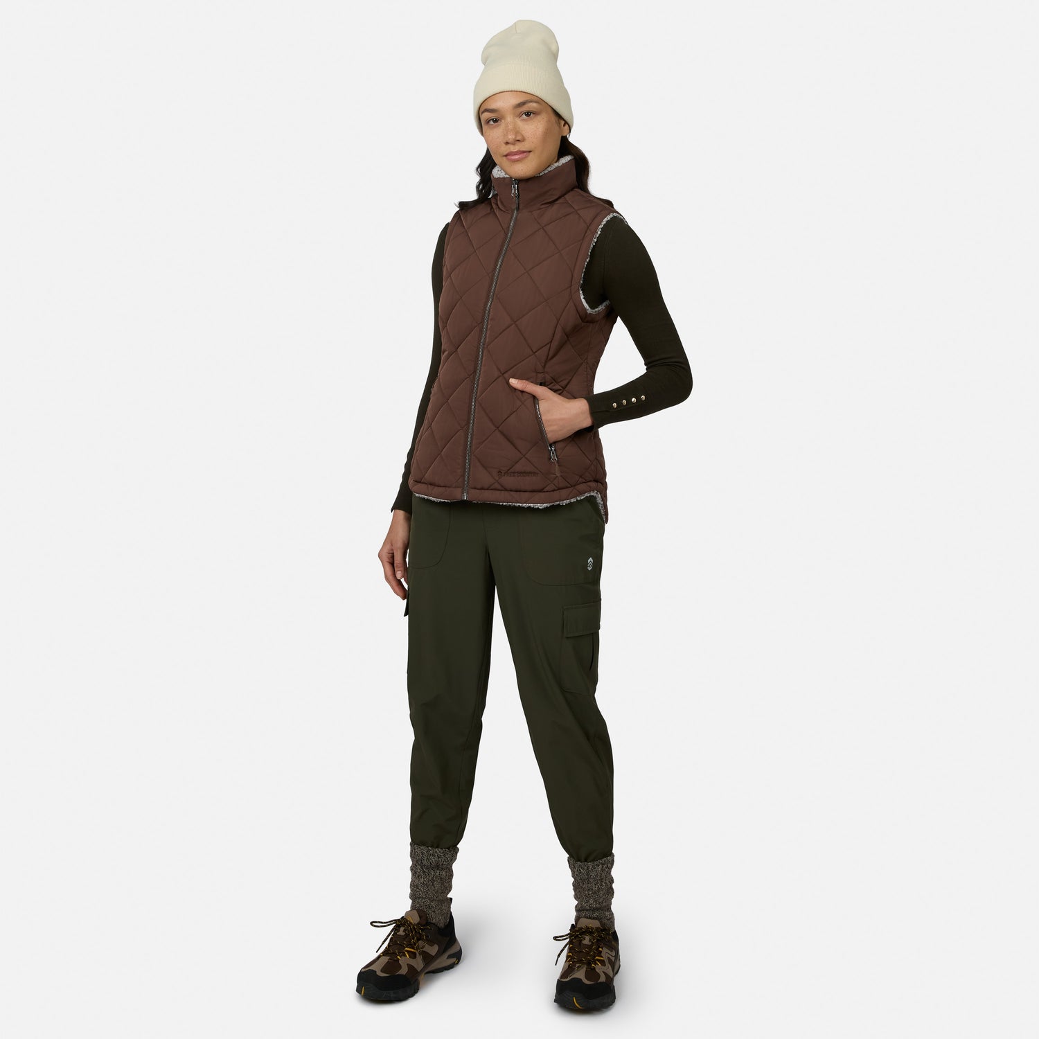 A woman stands against a plain white background wearing the Free Country Women’s Stratus Lite II Reversible Vest, crafted from recycled polyester in a brown quilted design. She pairs the lightweight, stylish, and sustainable vest with a dark long-sleeve shirt, green cargo pants, hiking shoes, gray socks, and a white beanie.