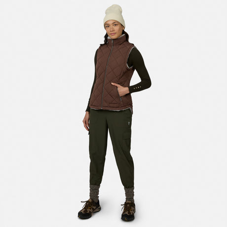 A woman stands against a plain white background wearing the Free Country Women’s Stratus Lite II Reversible Vest, crafted from recycled polyester in a brown quilted design. She pairs the lightweight, stylish, and sustainable vest with a dark long-sleeve shirt, green cargo pants, hiking shoes, gray socks, and a white beanie.