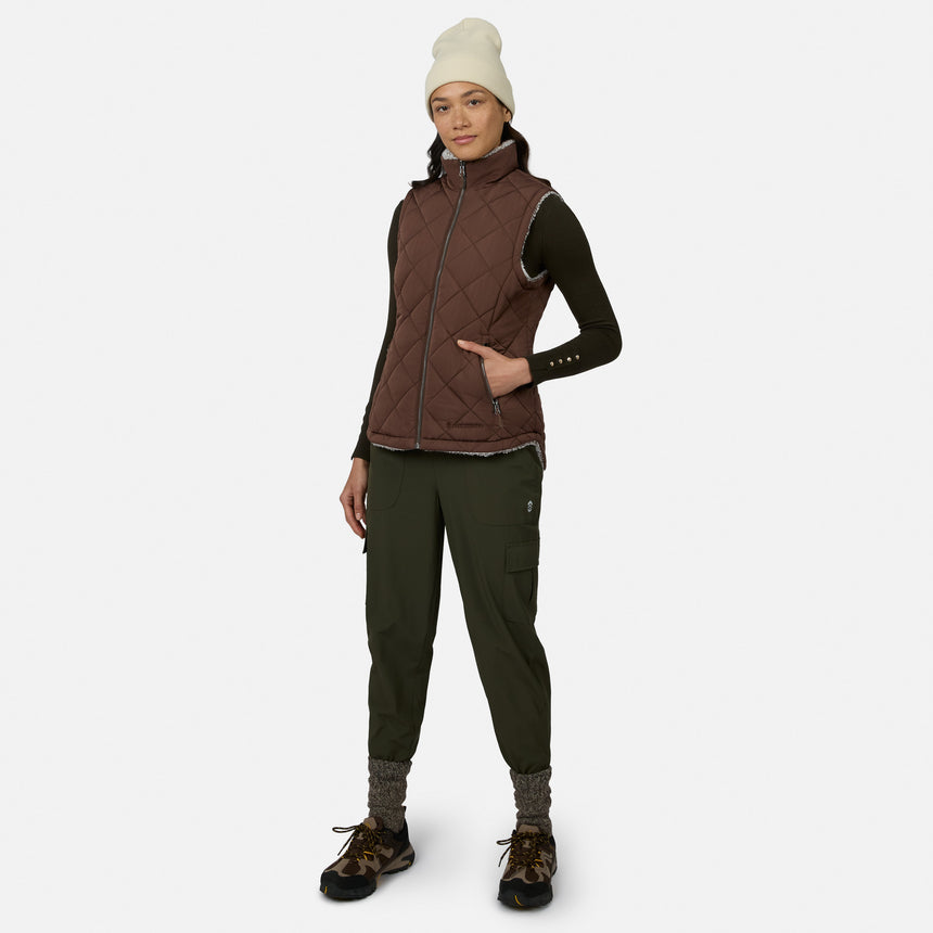 A woman stands against a plain white background wearing the Free Country Women’s Stratus Lite II Reversible Vest, crafted from recycled polyester in a brown quilted design. She pairs the lightweight, stylish, and sustainable vest with a dark long-sleeve shirt, green cargo pants, hiking shoes, gray socks, and a white beanie.