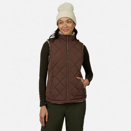 A woman stands smiling with her hand in her pocket against a plain white background, wearing a white beanie, a dark long-sleeve shirt, and the Free Country Women’s Stratus Lite II Reversible Vest in Sable—embodying lightweight warmth and effortless style.