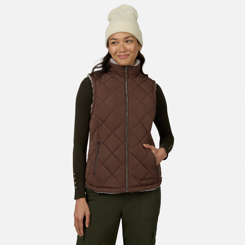 A woman stands smiling with her hand in her pocket against a plain white background, wearing a white beanie, a dark long-sleeve shirt, and the Free Country Women’s Stratus Lite II Reversible Vest in Sable—embodying lightweight warmth and effortless style.
