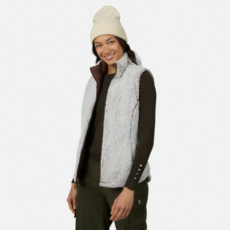 A smiling woman stands against a plain white background, effortlessly showcasing lightweight warmth and comfort in the Free Country Women’s Stratus Lite II Reversible Vest—a light gray sherpa vest made from recycled polyester—layered over a dark long-sleeve shirt, paired with a cream beanie and dark green pants.