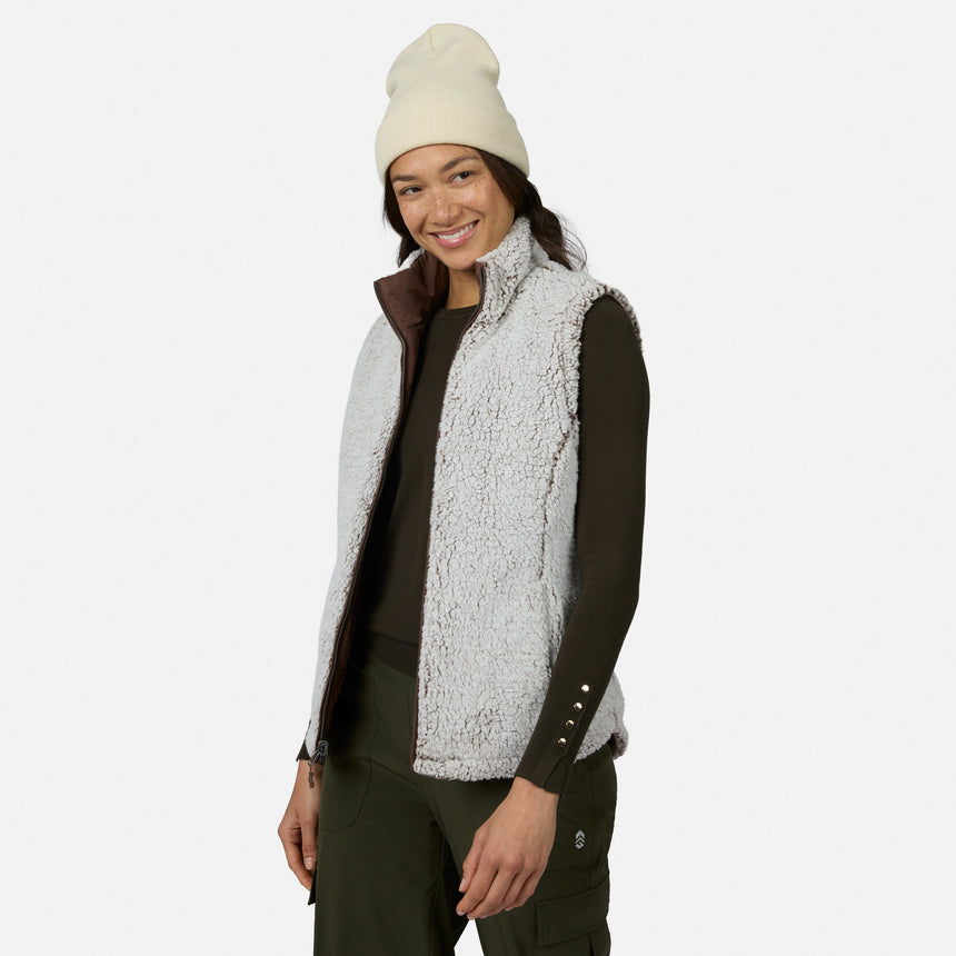 A smiling woman stands against a plain white background, effortlessly showcasing lightweight warmth and comfort in the Free Country Women’s Stratus Lite II Reversible Vest—a light gray sherpa vest made from recycled polyester—layered over a dark long-sleeve shirt, paired with a cream beanie and dark green pants.