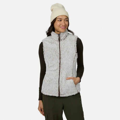 A woman stands against a plain white background, smiling and looking to the side while wearing the Free Country Women’s Stratus Lite II Reversible Vest in white—crafted from recycled polyester—layered over a black long-sleeve shirt, with olive green pants and a white beanie. This versatile vest delivers lightweight warmth and sustainable style.