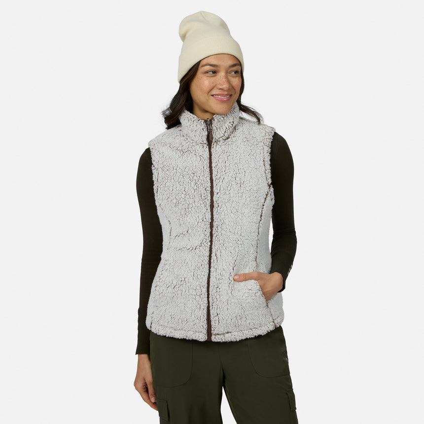 A woman stands against a plain white background, smiling and looking to the side while wearing the Free Country Women’s Stratus Lite II Reversible Vest in white—crafted from recycled polyester—layered over a black long-sleeve shirt, with olive green pants and a white beanie. This versatile vest delivers lightweight warmth and sustainable style.