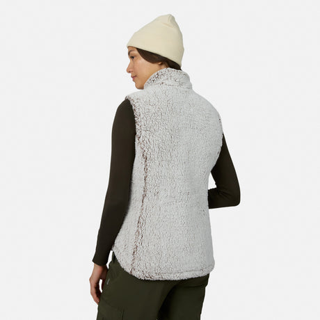 A woman stands facing away, looking over her shoulder against a plain white background while wearing a cream beanie, dark long-sleeve shirt, olive green pants, and the Free Country Women’s Stratus Lite II Reversible Vest in light gray sherpa fleece made from recycled polyester. This stylish vest delivers lightweight warmth with an eco-friendly touch.