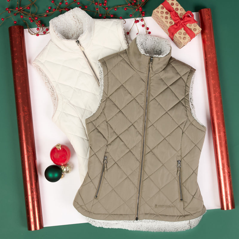 Two quilted sleeveless puffer vests with faux-fur trim are arranged on a white sheet of wrapping paper with holiday decorations. One vest is cream/off-white and the other is taupe-beige, both with diagonal quilting and front zippers.