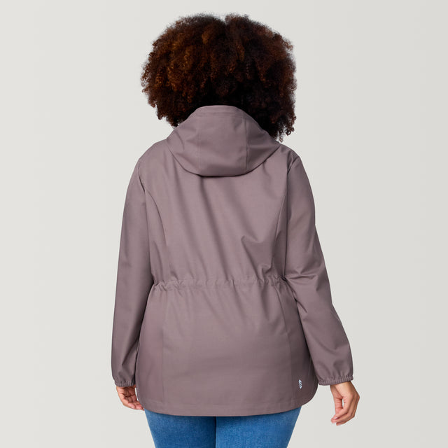 A person with curly hair is seen from behind wearing the Free Country Women's Plus Size X2O Anorak Rain Jacket in mauve and blue jeans, standing against a plain light background.