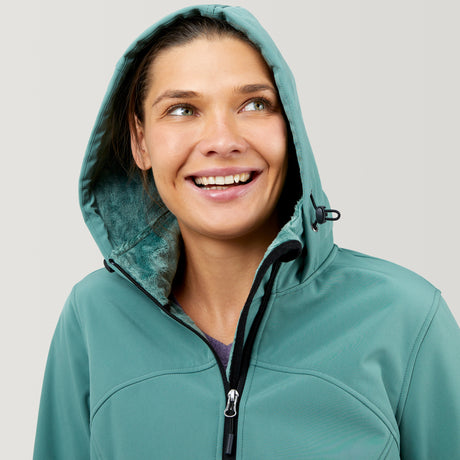 A smiling woman wears the Women's FreeCycle® Super Softshell® Jacket by Clearance in teal, zipped up with the hood on. She looks up and to the side against a plain light background. The jacket is water resistant and softshell.