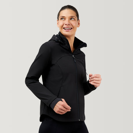 A woman models the Clearance Women's FreeCycle® Super Softshell® Jacket in black, standing against a light background. She smiles and looks to the side with one hand raised slightly, showing off the jacket's hooded design.
