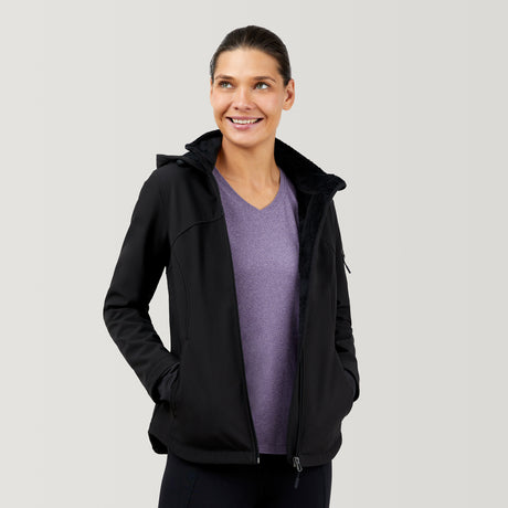 A woman wearing the Clearance Women's FreeCycle® Super Softshell® Jacket in black over a purple V-neck shirt stands with her hands in her pockets, smiling and looking to the side against a plain, light-colored background.