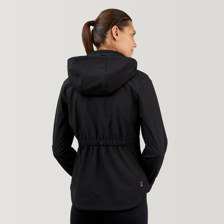 A woman with brown hair in a ponytail wears the Clearance Women's FreeCycle® Super Softshell® Jacket, featuring an elastic waistband and long sleeves. She faces away, highlighting its four-way stretch design against a plain light background.