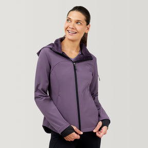 A woman with dark hair pulled back smiles and looks to the side, wearing the Clearance Women's FreeCycle® Super Softshell® Jacket with a hood, against a plain light background. She rests her hands on the water-resistant jacket.