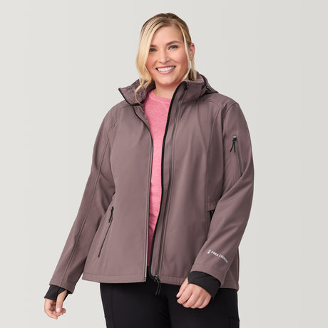 [Lola is 5'10.5" wearing a size 1X.] Woman wearing the Free Country Women's Plus Size Aeris II Super Softshell® Jacket in brown, featuring metallic zippers, a detachable hood, and storm cuffs, smiling with hands in pockets against a light-colored background.
