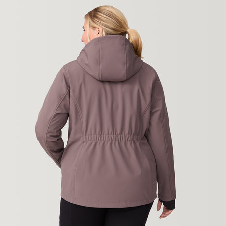 [Lola is 5'10.5" wearing a size 1X.] Woman wearing the Free Country Women's Plus Size Aeris II Super Softshell® Jacket in brown, featuring metallic zippers, a detachable hood, and storm cuffs, smiling with hands in pockets against a light-colored background.