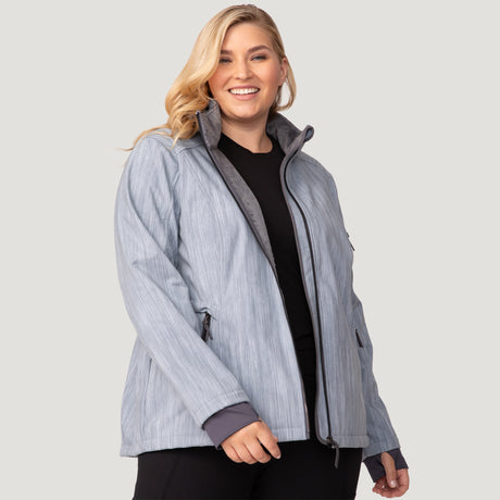 A smiling woman with long blonde hair models the Free Country Women's Plus Size Aeris II Super Softshell® Jacket in light gray over a black top and pants, standing against a plain light gray background.