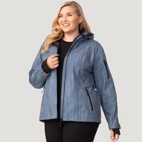 A smiling woman with long blonde hair wears the Free Country Women's Plus Size Aeris II Super Softshell® Jacket, featuring water-resistant three-layer bonded fabric, over a black top and pants, standing against a light gray background.