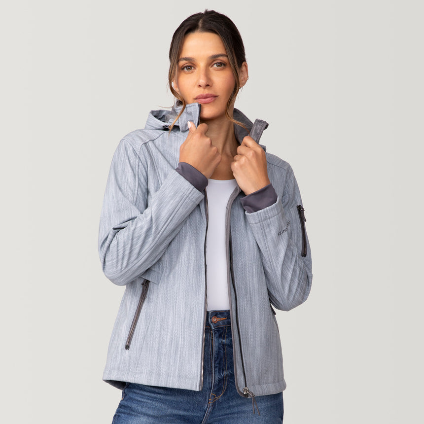 A woman wearing a light blue Free Country Women’s Aeris II Super Softshell® Jacket, featuring a chevron pattern, a zip-up hood, and dark gray accents.