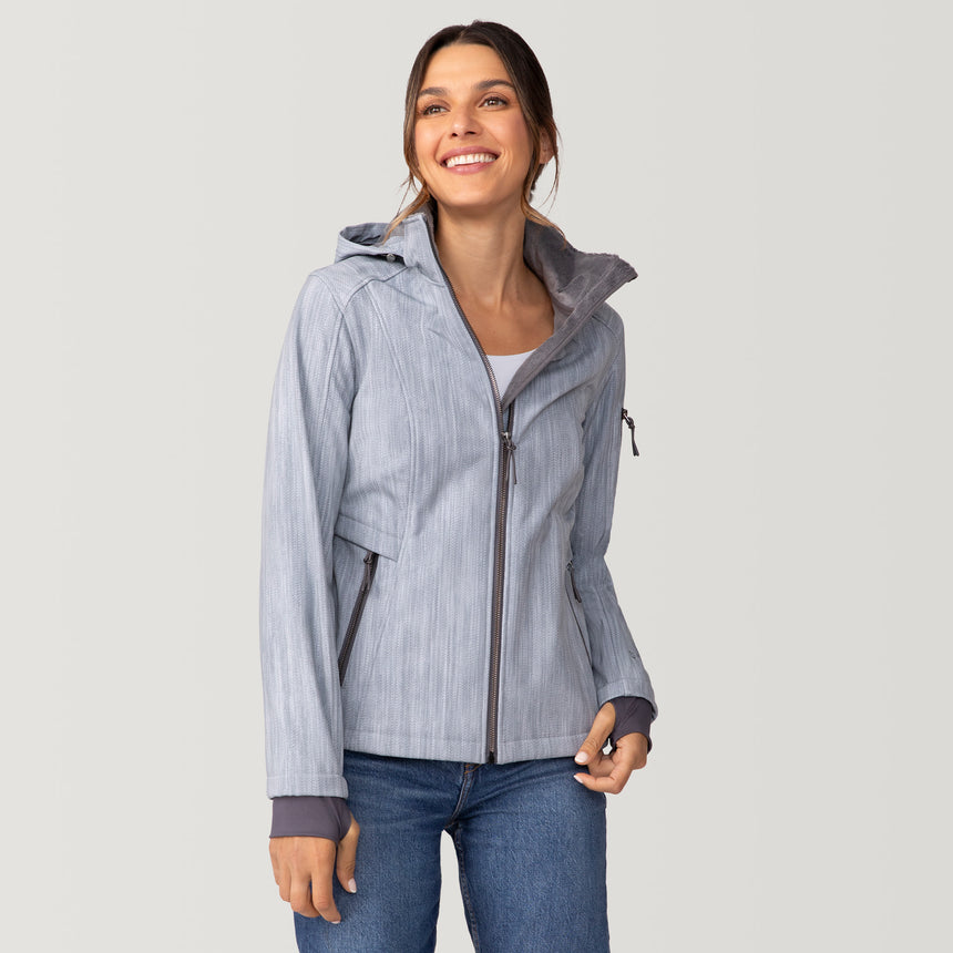 A smiling woman wears a grey, water-resistant Women’s Aeris II Super Softshell® Jacket with a chevron pattern, dark grey cuffs, and a zippered front over blue jeans.
