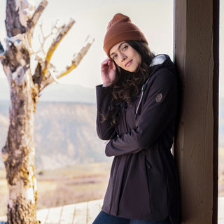 Woman in a purple coat and brown beanie leaning against a wall with a scenic background