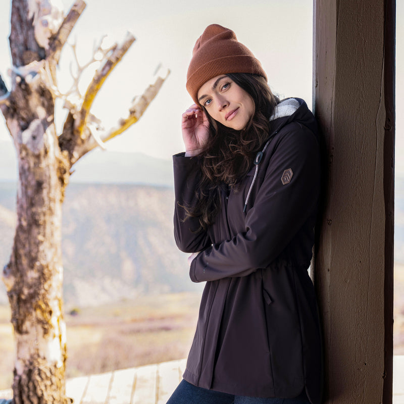 Woman in a purple coat and brown beanie leaning against a wall with a scenic background