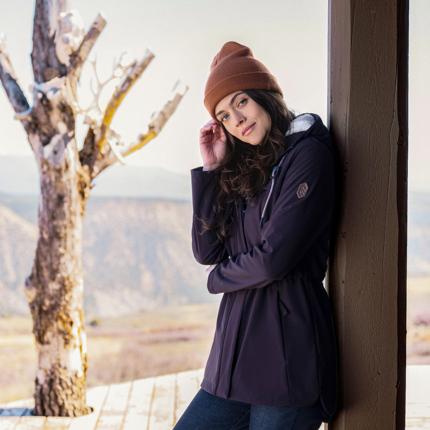 Woman in a purple coat and brown beanie leaning against a wall with a scenic background
