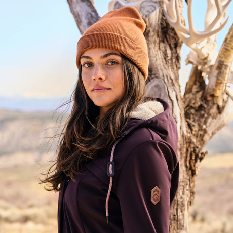 Woman wearing a brown beanie and purple jacket standing next to a tree in a desert-like setting