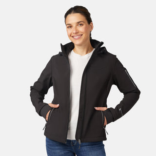 A smiling woman wears the Free Country Women's StormTech Super Softshell® Jacket in black over a white shirt and blue jeans, standing with hands in her jacket pockets against a plain light background.