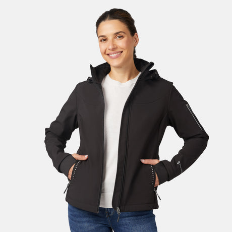A smiling woman wears the Free Country Women's StormTech Super Softshell® Jacket in black over a white shirt and blue jeans, standing with hands in her jacket pockets against a plain light background.